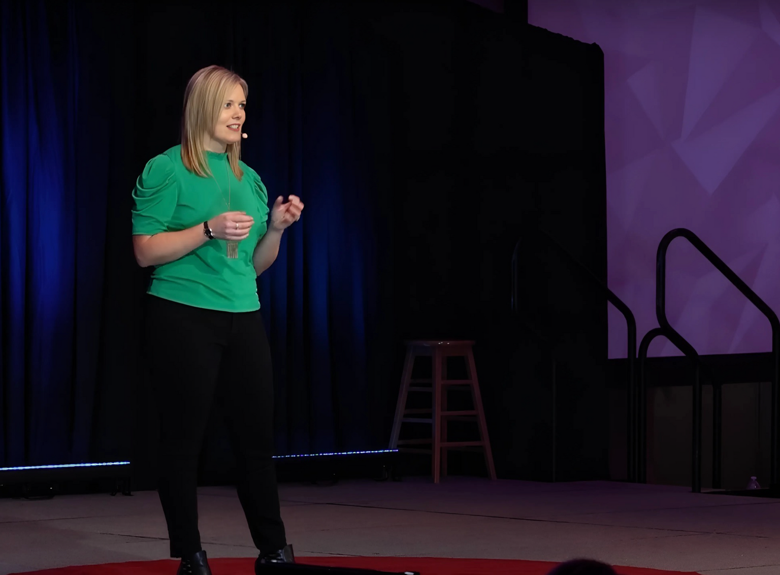 Woman in green shirt and black pants standing on a stage with a purple backdrop